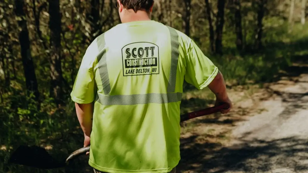 Scott Construction worker wearing neon yellow safety vest with company logo and Lake Delton, Wisconsin branding, working on residential driveway surrounded by green vegetation and trees.